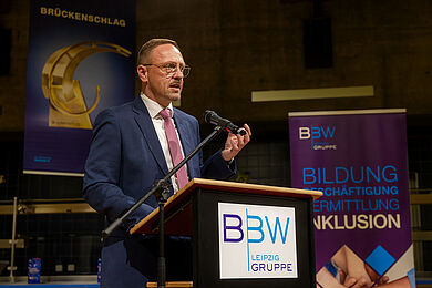 Tobias Schmidt am Rednerpult. Vorne das logo der BBW-Leipzig-Gruppe. Er guckt ernst und gestikuliert mit seiner linken Hand. Er trägt einen dunklen Anzug über einem weißen Hemd mit einer rot-weißen Krawatte. Rechts steht das Rollup der BBW-Leipzig-Gruppe auf dem steht: Bildung, Beschäftigung, Vermittlung, Inklusion