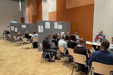 In einem großen Raum sind Bereiche mit Stellwänden unterteilt. In den Abteilen sitzen mehrere Menschen im Kreis für die Lebendige Bibliothek