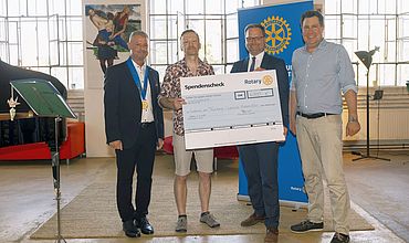 Vier Männer stehen nebeneinander in einem hellen Raum mit großen Fenstern. Zwei von ihnen halten gemeinsam einen übergroßen Spendenscheck des Rotary Clubs in der Hand. Auf dem Scheck ist ein Betrag von 6.000 Euro zu sehen, bestimmt für ein Projekt namens „Diakonie am Thonberg – Inklusive Klassenfahrt“. Im Hintergrund sind ein Banner des Rotary Clubs, moderne Kunstwerke sowie Pflanzen und Sitzmöbel zu erkennen. Die Stimmung wirkt freundlich und formell.