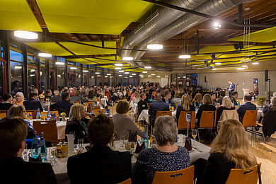 Blick in die Mensa des BBW. An langen Tischen sitzen zahlreiche Gäste. Sie blicken alle nach vorne auf die Bühne, auf der man einen Redner erkennen kann.