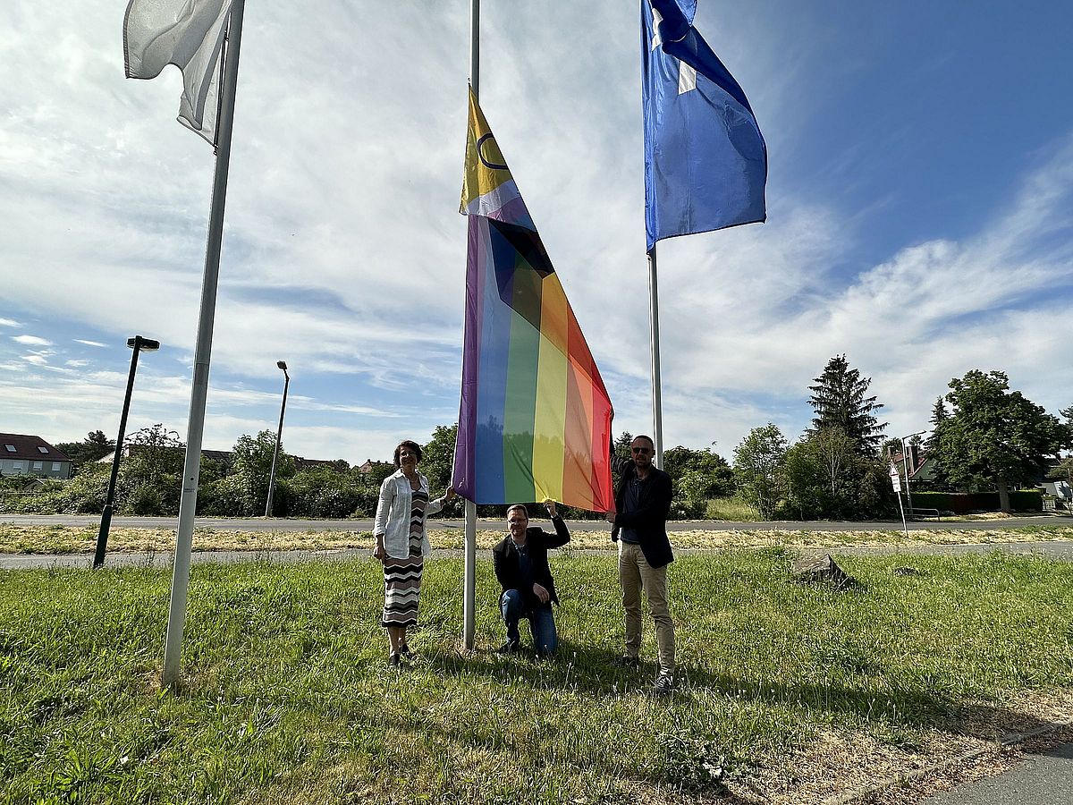 Auf einer Wiese vor blauem Himmel hängen drei Fahnen, in der Mitte die Progress Pride Flag. Unter der Flagge stehen von links nach rechts: Dr. Grit Franke, Geschäftsbereichsleiterin BBW, Tobias Schmidt, Hauptgeschäftsführer BBW-Leipzig-Gruppe und Bastian Schmiedel, Kaufmännischer Geschäftsführer BBW-Leipzig-Gruppe.