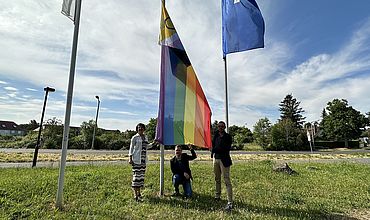 Auf einer Wiese vor blauem Himmel hängen drei Fahnen, in der Mitte die Progress Pride Flag. Unter der Flagge stehen von links nach rechts: Dr. Grit Franke, Geschäftsbereichsleiterin BBW, Tobias Schmidt, Hauptgeschäftsführer BBW-Leipzig-Gruppe und Bastian Schmiedel, Kaufmännischer Geschäftsführer BBW-Leipzig-Gruppe.